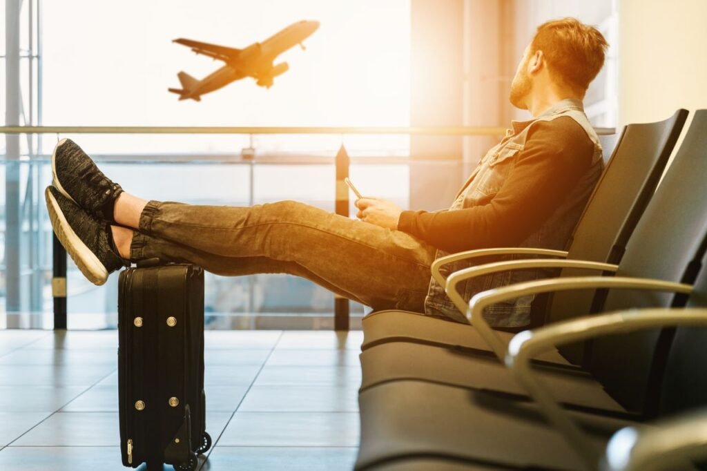 Side view full body of bearded man sitting on chair in waiting hall of airport and looking at flying aircraft behind window